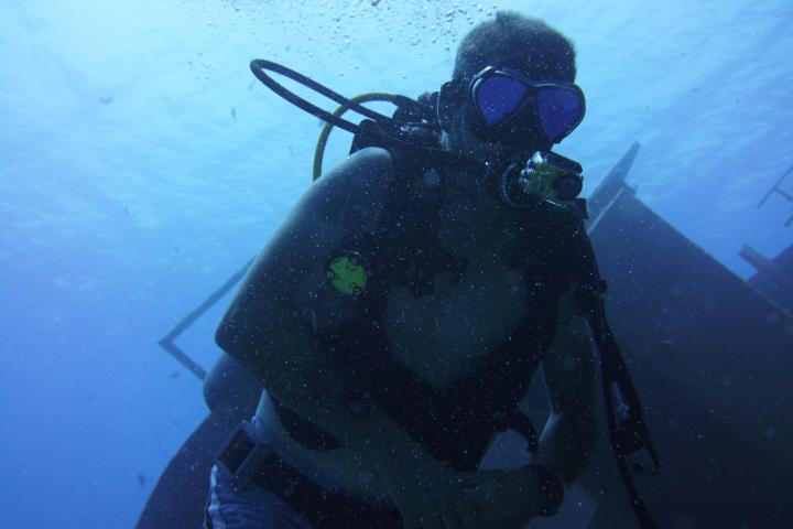 Scuba diver underwater near a sunken ship, surrounded by clear blue water.