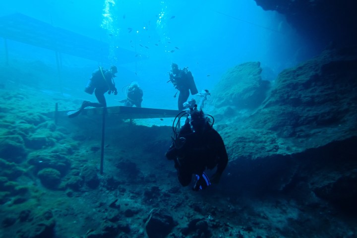 Four scuba divers underwater near a rocky cave.