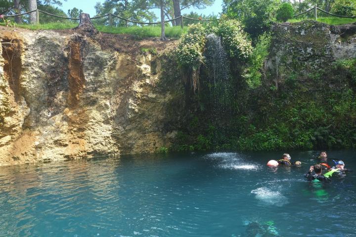 Divers in a turquoise pool beneath a rocky cliff with greenery and a waterfall.