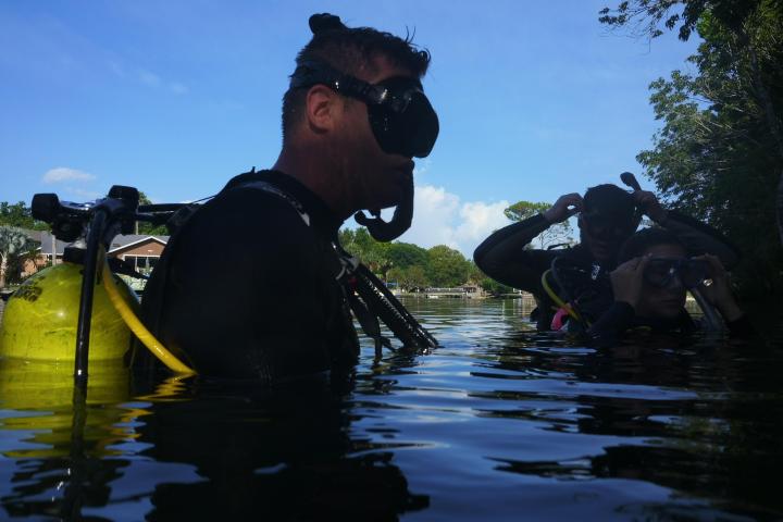 Three scuba divers adjusting gear in water near trees and houses.