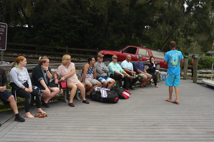 Group of people sitting on a dock, with a person in blue standing, next to a parked red SUV and a park sign.
