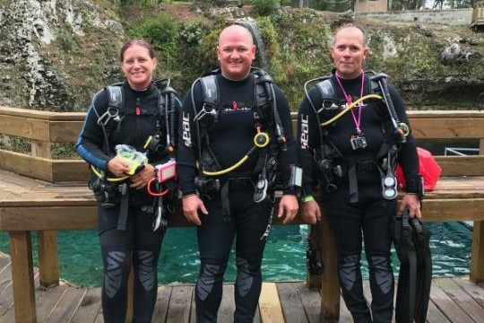 Three scuba divers in wetsuits stand on a wooden deck beside a body of water.