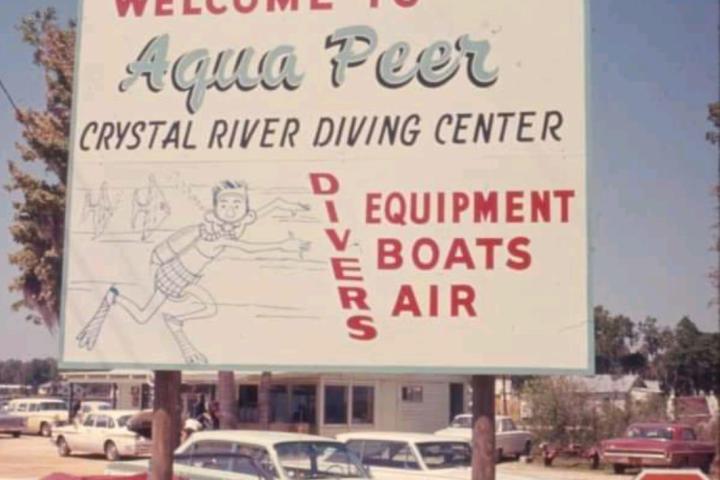 Vintage sign for Aqua Peer dive shop with cars and a building in the background.