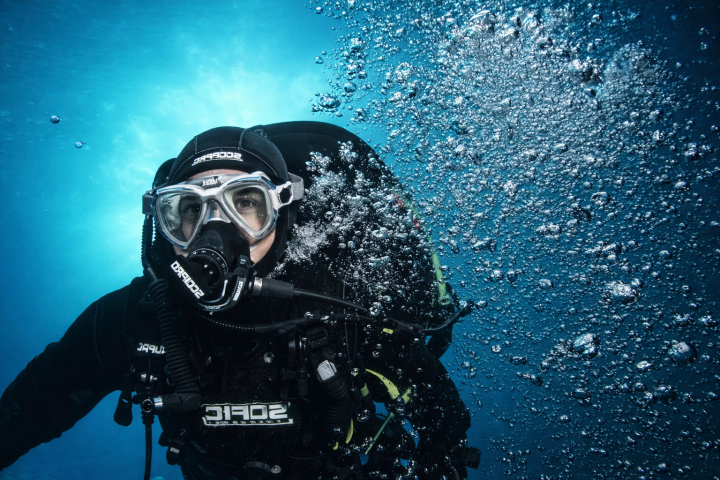 Diver underwater with scuba gear, surrounded by bubbles in clear blue water.