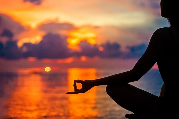 Silhouette of a person meditating by the water at sunset, with clouds and vibrant orange sky.