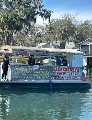 Pontoon boat with 'Sea Daddys Dive Center' ad, people onboard, trees and houses in the background.