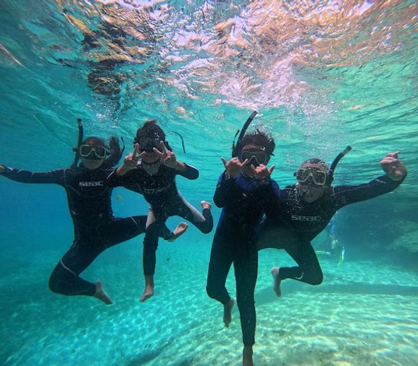 Four snorkelers underwater making hand gestures and wearing black wetsuits.