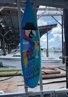 Surfboard with a parrot holding a drink, text 'It's 5 o'clock somewhere,' dock and boats in the background.